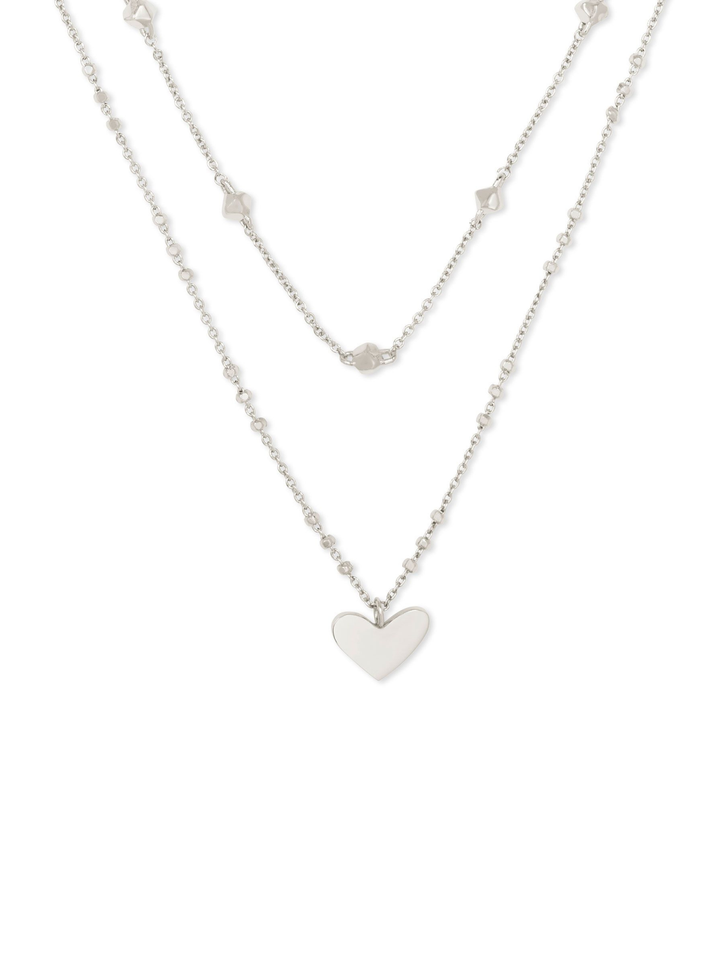 A silver double-layered necklace featuring a delicate chain with small star-shaped charms and a longer chain with a polished heart pendant, displayed against a solid white background.