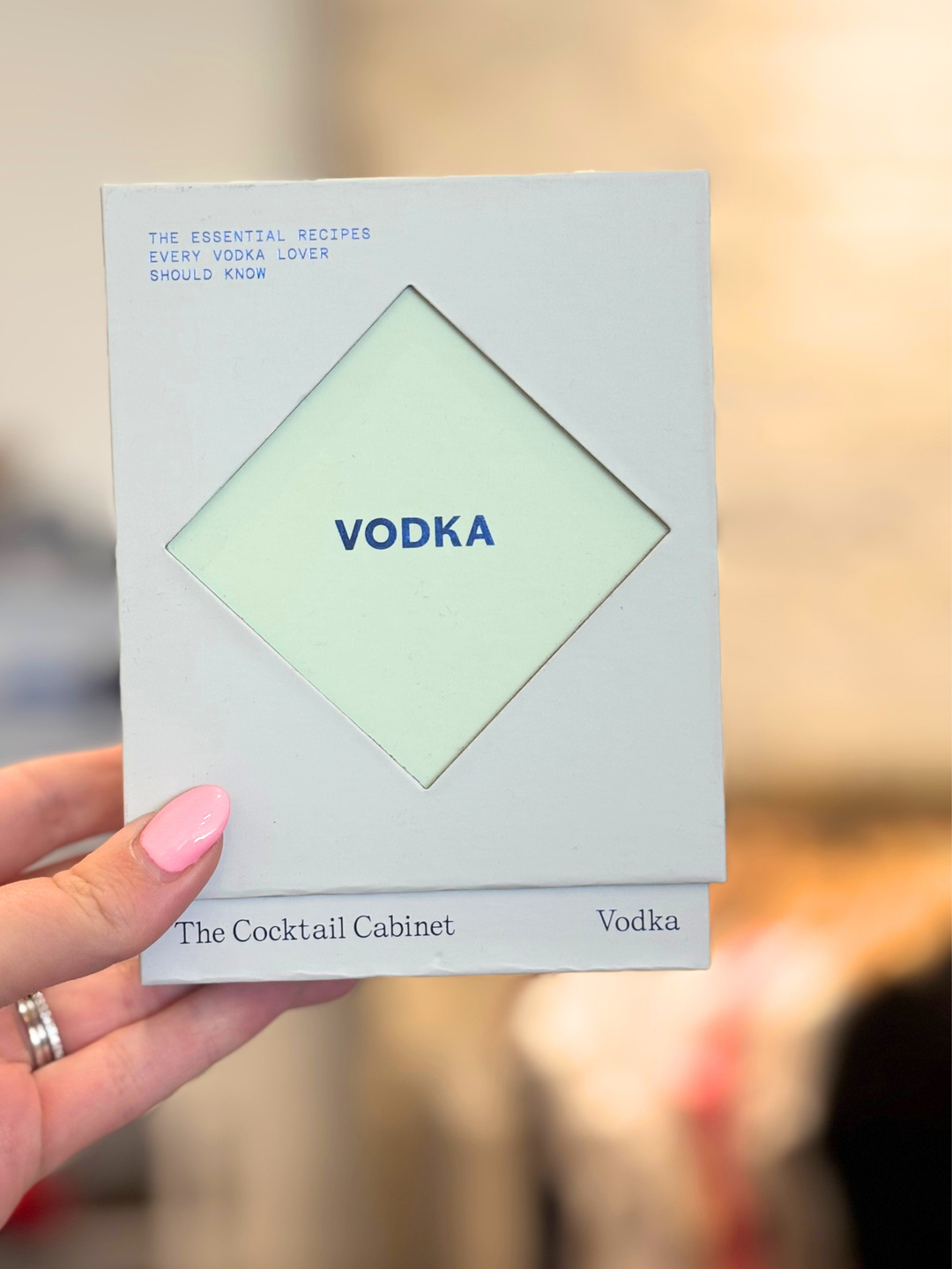 A hand with pink nail polish holds a square white card or booklet featuring a pale green diamond shape in the center with the word &