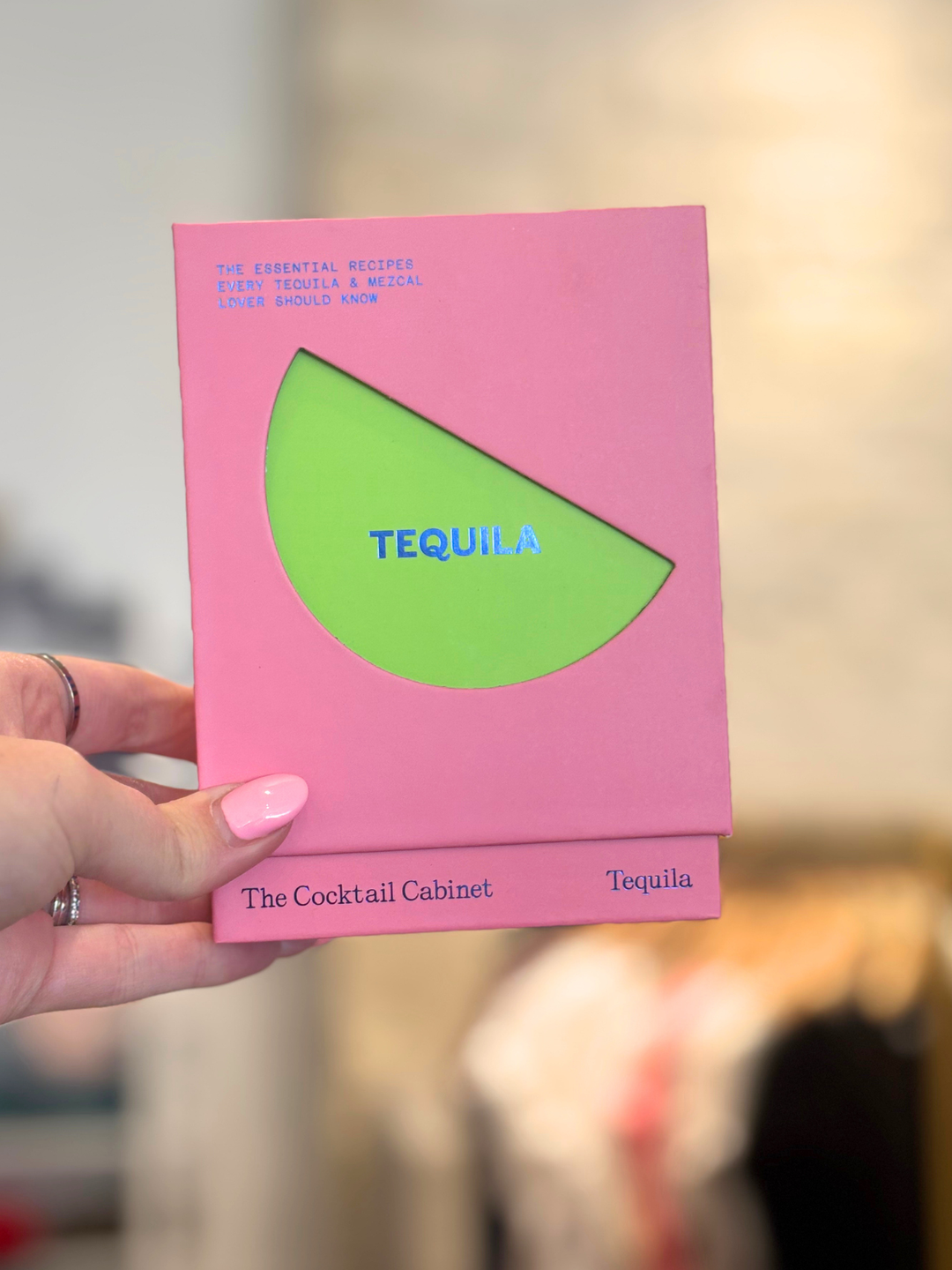 A hand with pink nail polish holds a pink cocktail cabinet box with a green semicircle cutout and the word &