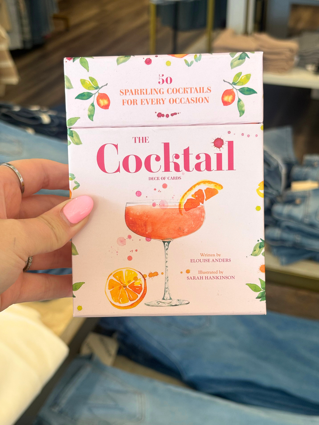 A hand with pink nail polish holds a book titled "The Cocktail" in front of a blurred background of blue jeans and clothing racks. The book cover is light pink with illustrations of orange slices, green leaves, and a pink cocktail glass.