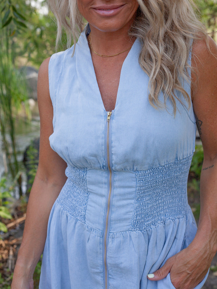 A woman wearing a light blue sleeveless dress with a smocked waist and a front zipper, photographed outdoors with greenery in the background.