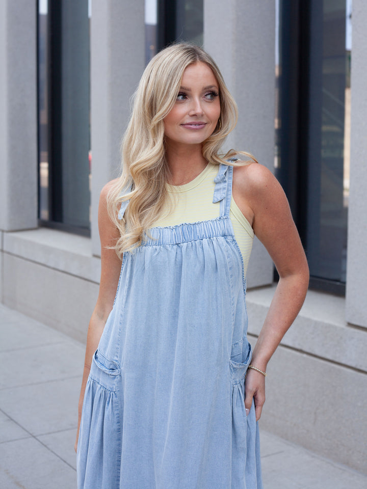 A blonde woman wearing a light blue denim overall dress over a pale yellow tank top, standing outdoors in front of a modern building with gray columns.