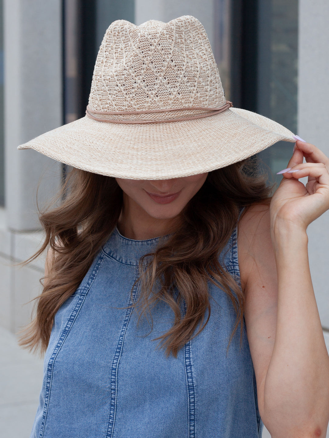 A woman wearing a light beige woven sun hat with a wide brim and a blue denim sleeveless top, photographed outdoors against a blurred gray building background.