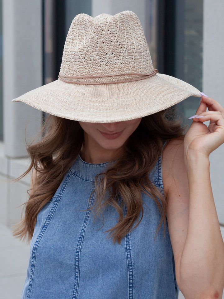 A woman wearing a light beige woven sun hat with a wide brim and a blue denim sleeveless top, photographed outdoors against a blurred gray building background.