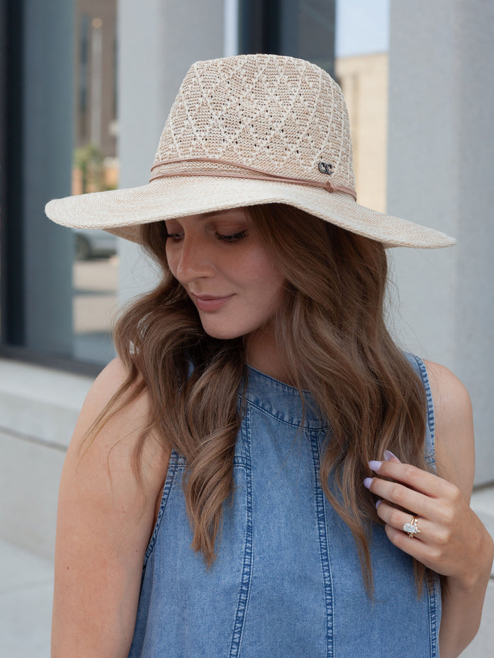 A woman wearing a wide-brimmed straw hat with a textured woven crown and a blue denim sleeveless top, photographed outdoors in front of a modern building.