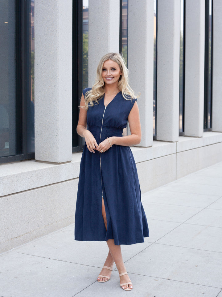 A woman wearing a sleeveless navy blue midi dress with a front zipper and side slit, standing outdoors in front of a modern building with large columns.