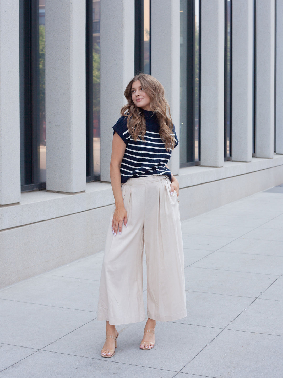 A woman stands outdoors in front of a modern building with large gray columns, wearing a navy and white horizontally striped short-sleeve top with cream wide-leg cropped pants and nude heels.