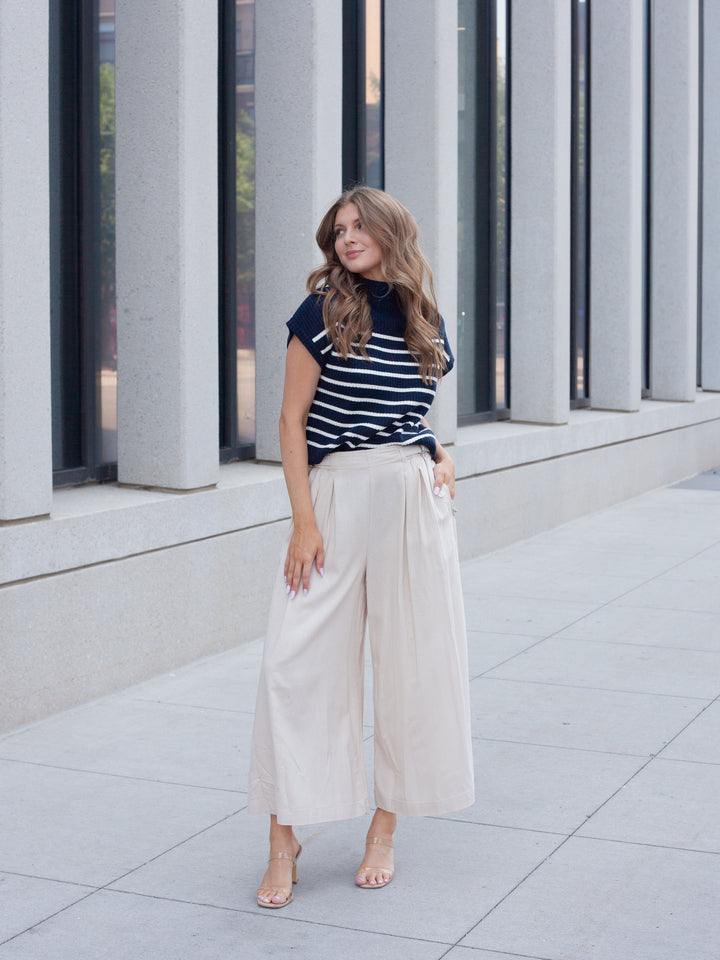 A woman stands outdoors in front of a modern building with large gray columns, wearing a navy and white horizontally striped short-sleeve top with cream wide-leg cropped pants and nude heels.