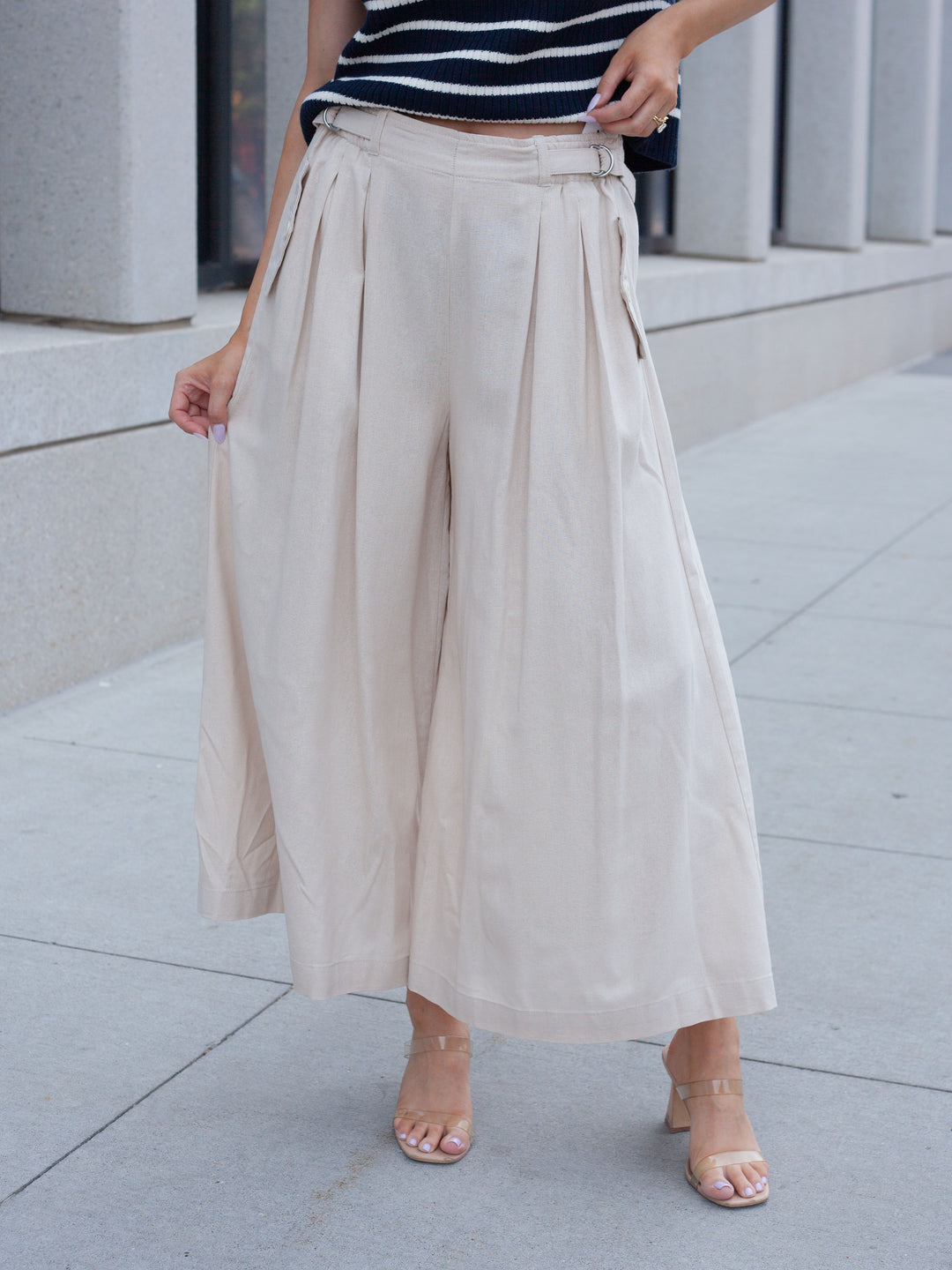 A person wearing high-waisted, wide-leg cropped pants in a light beige or cream color, paired with a navy and white striped top and clear-strap heeled sandals, standing outdoors on a paved walkway.
