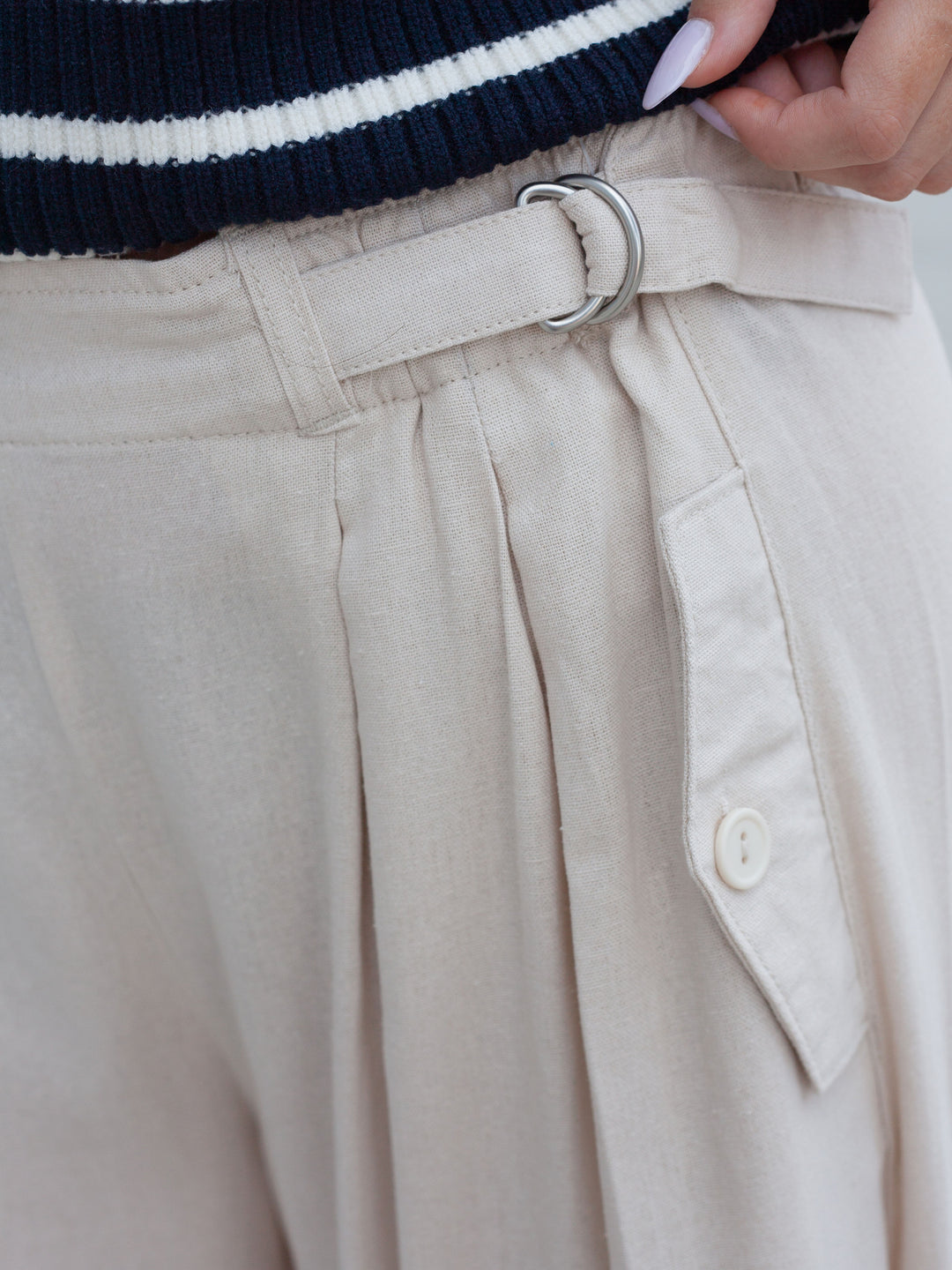 Close-up of a person wearing light beige pleated trousers with a matching belt and silver O-ring, paired with a navy and white striped sweater.