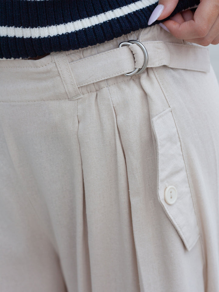 Close-up of a person wearing light beige pleated trousers with a matching belt and silver O-ring, paired with a navy and white striped sweater.