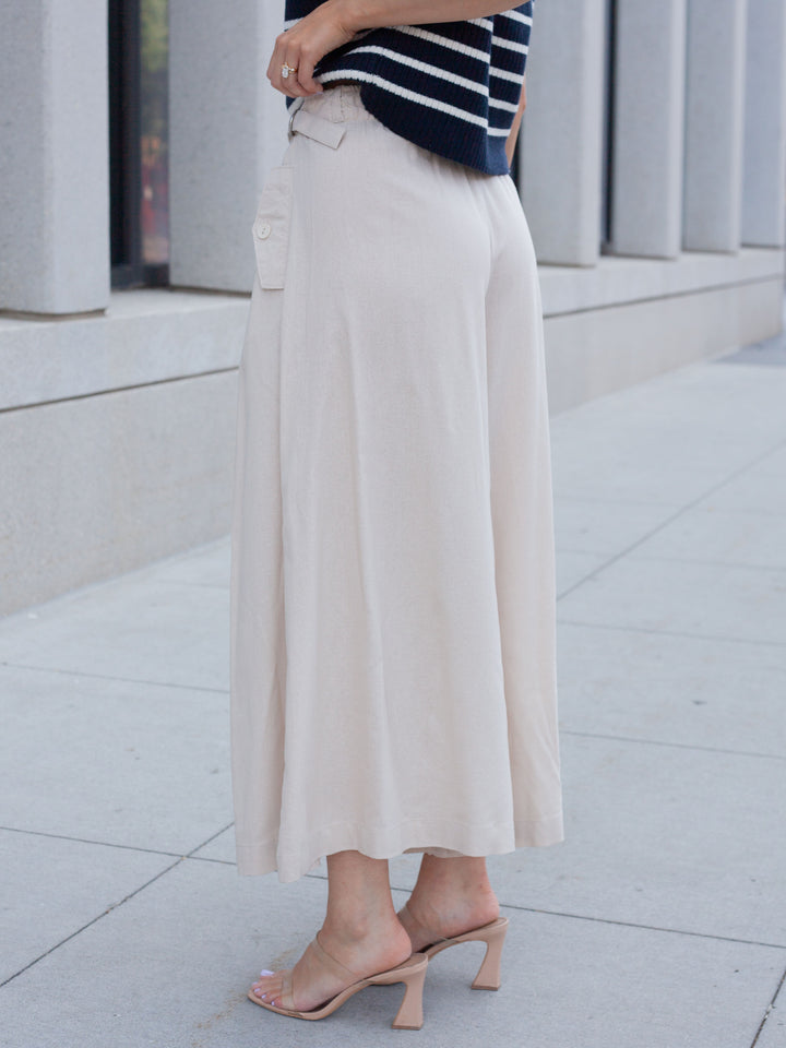 A person wearing a long cream-colored midi skirt with side flap pockets, paired with a navy and white striped top and nude heeled sandals, standing outdoors on a light gray paved walkway.