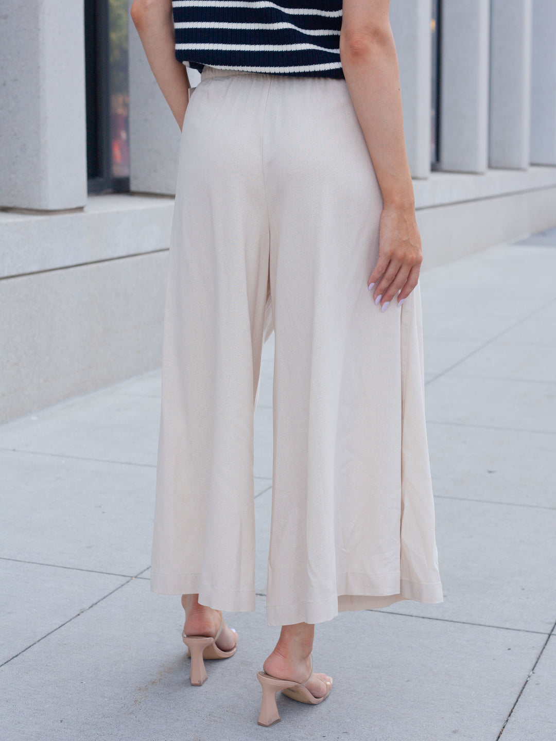 A person shown from the back wearing high-waisted, wide-leg cropped pants in a light cream or off-white color, paired with a navy and white striped top and nude heeled sandals, standing on a paved outdoor walkway.