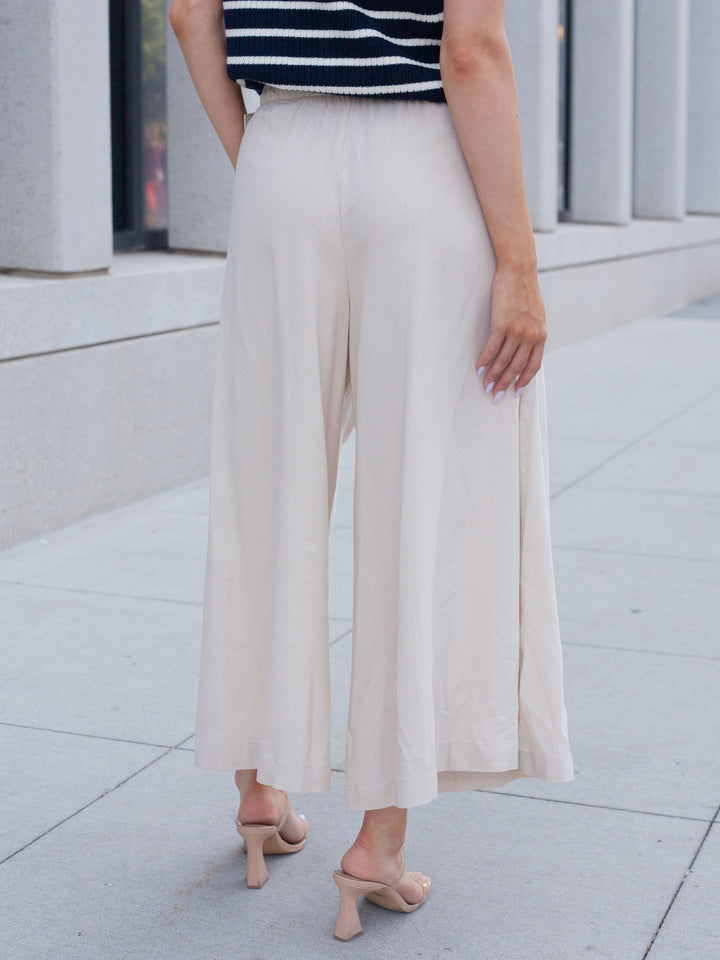 A person shown from the back wearing high-waisted, wide-leg cropped pants in a light cream or off-white color, paired with a navy and white striped top and nude heeled sandals, standing on a paved outdoor walkway.