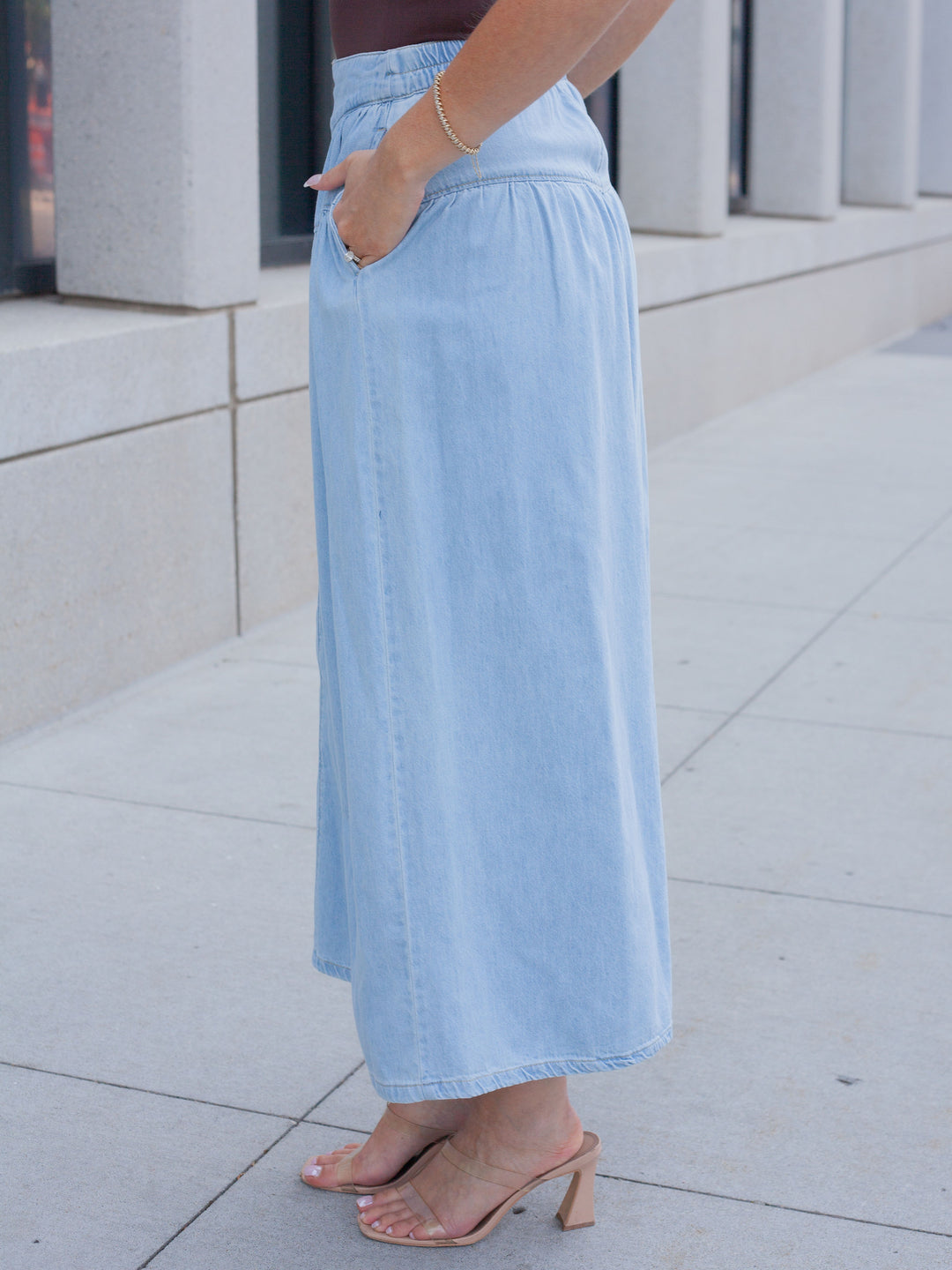 A person wearing a light blue denim maxi skirt with side pockets, paired with nude heeled mule sandals, standing outdoors on a paved walkway.