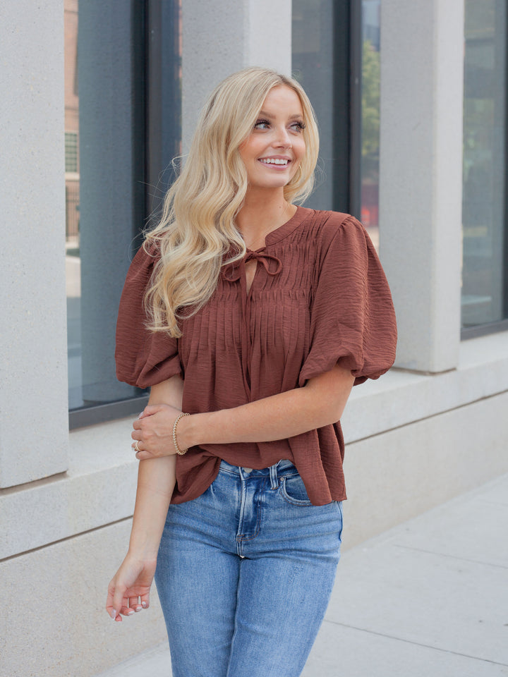 A woman with long blonde hair wearing a rust-brown crinkled puff-sleeve blouse and blue jeans, standing outdoors in front of a modern building with large windows.