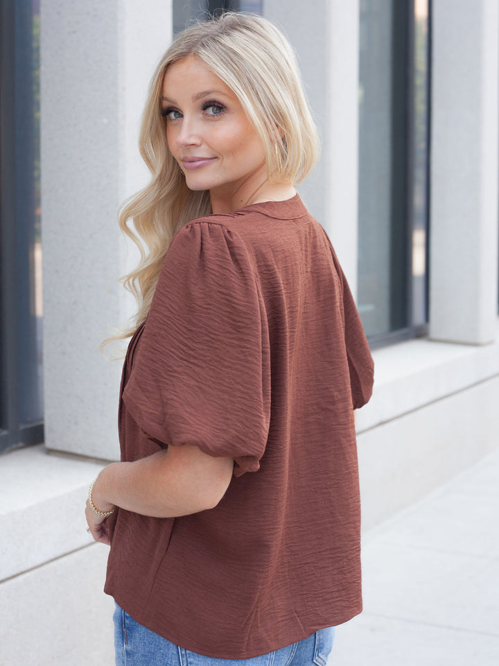 A woman with blonde hair wearing a textured brown puff-sleeve top and blue jeans, photographed from the back in an outdoor setting with a modern building facade in the background.