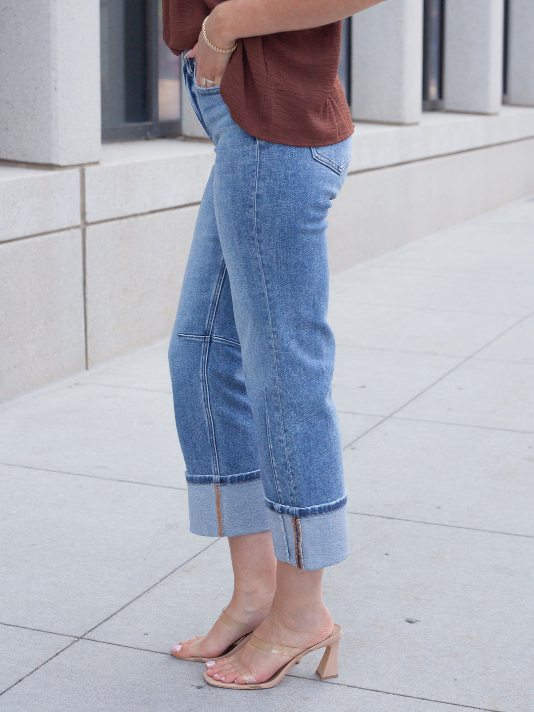 A person wearing light blue cropped denim jeans with rolled cuffs and side zipper details, paired with a brown top and clear heeled sandals, standing on a light concrete sidewalk.