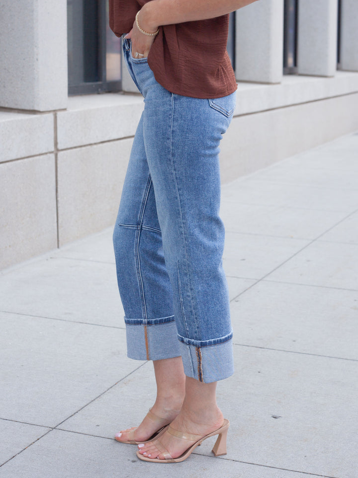 A person wearing light blue cropped denim jeans with rolled cuffs and side zipper details, paired with a brown top and clear heeled sandals, standing on a light concrete sidewalk.