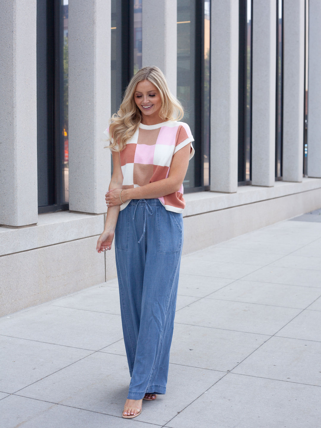 A woman stands outdoors on a paved walkway in front of a modern building with large columns. She is wearing a short-sleeve cropped knit top with a pink, white, and light peach checkerboard pattern, paired with wide-leg blue jeans.