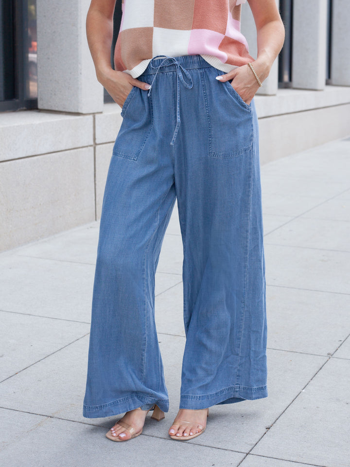 A person wearing wide-leg blue denim pants with a drawstring waist, paired with a pink, white, and beige checkered top and clear-strap sandals, standing on a light gray tiled sidewalk.