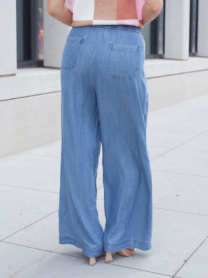 A person shown from the back wearing light blue wide-leg denim pants with back patch pockets, standing barefoot on a light concrete sidewalk.