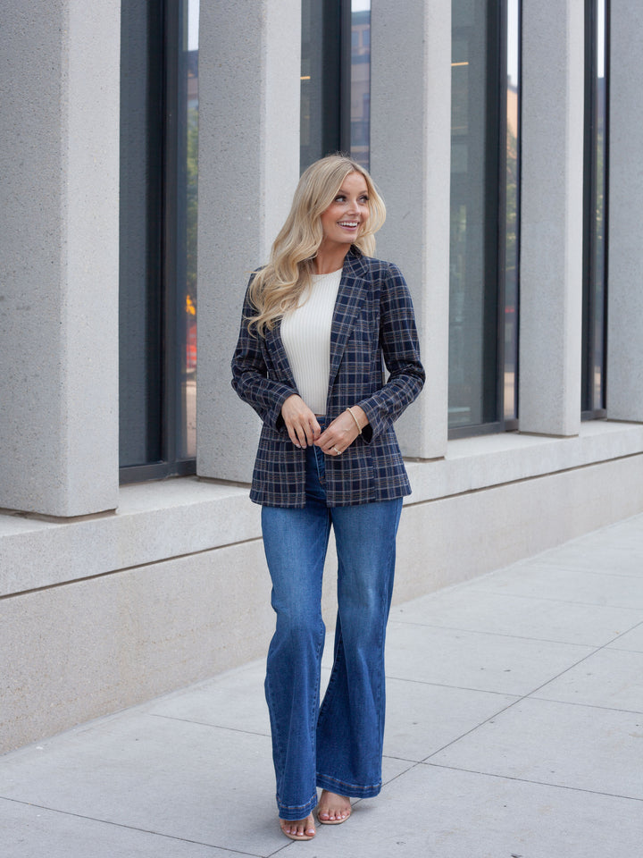 A woman stands outdoors on a sidewalk in front of a modern building with large gray columns and dark windows. She is wearing a navy and black plaid blazer over a white top, blue flare jeans, and is barefoot.