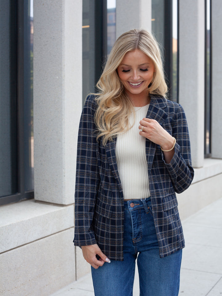 A woman wearing a navy blue plaid blazer over a cream ribbed top and blue jeans, standing outdoors in front of a modern building with large windows and concrete columns.