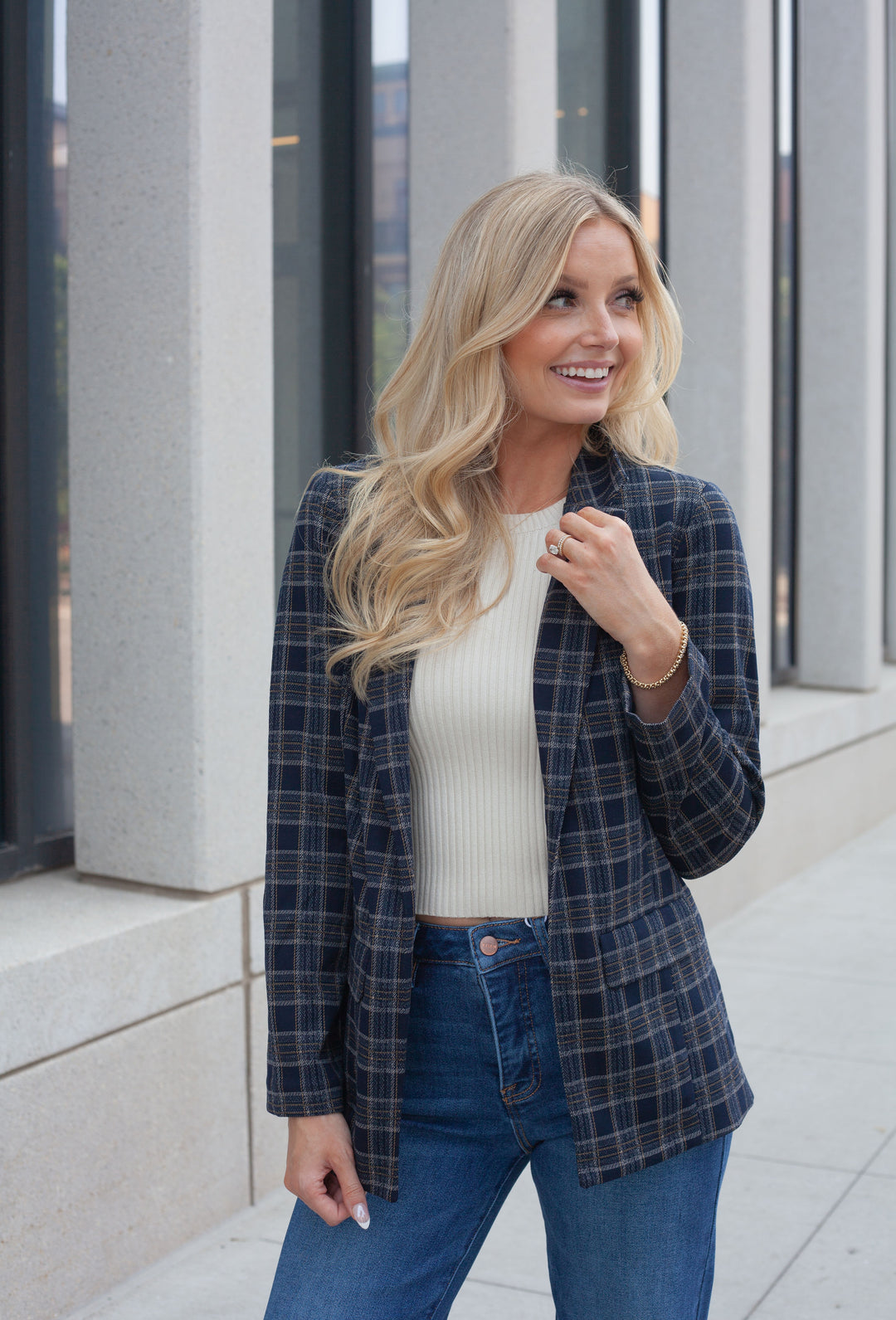 A woman with blonde hair stands outdoors wearing a navy and gray plaid blazer over a cream ribbed top and blue jeans.