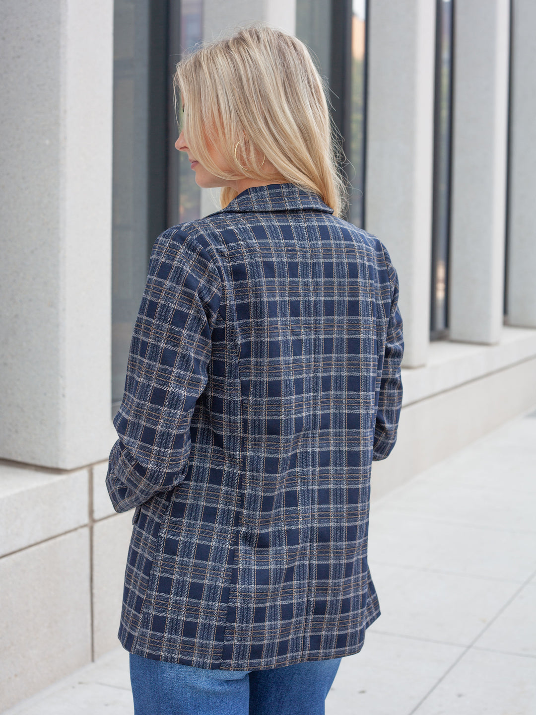 A woman with blonde hair is shown from behind wearing a navy blue plaid button-up shirt with white and gray lines, paired with blue jeans, standing outdoors against a modern building facade.