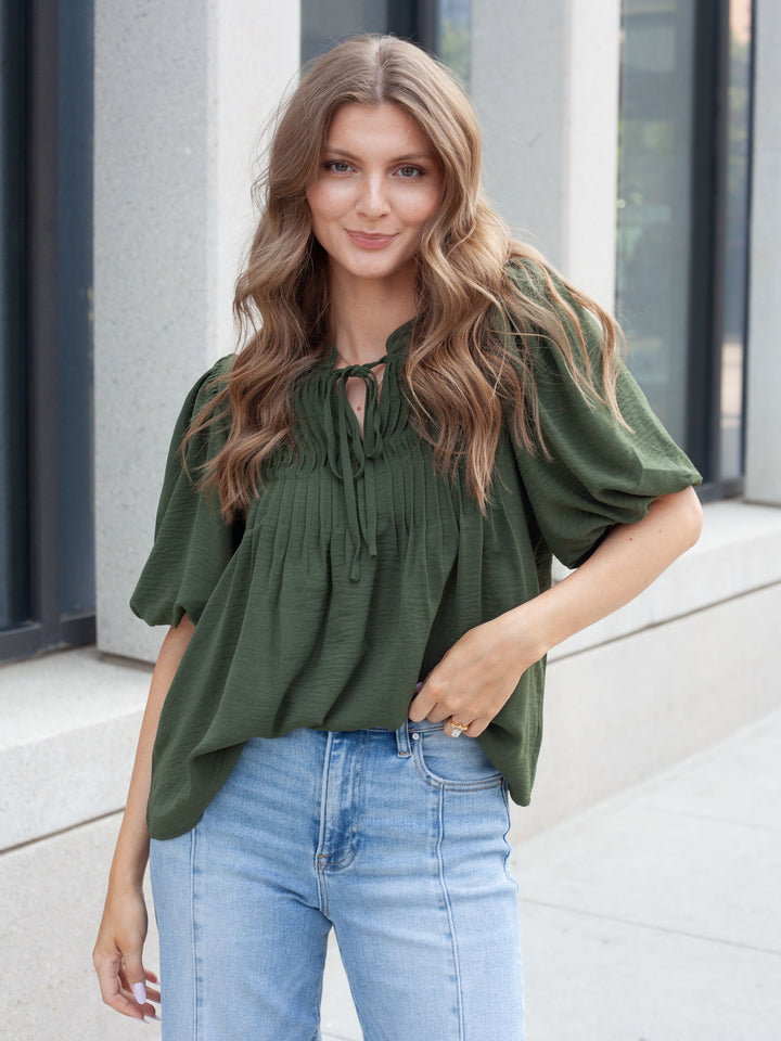 A woman wearing an olive green short-sleeve blouse with a tie-neck detail and light blue jeans, standing outdoors against a modern building facade.