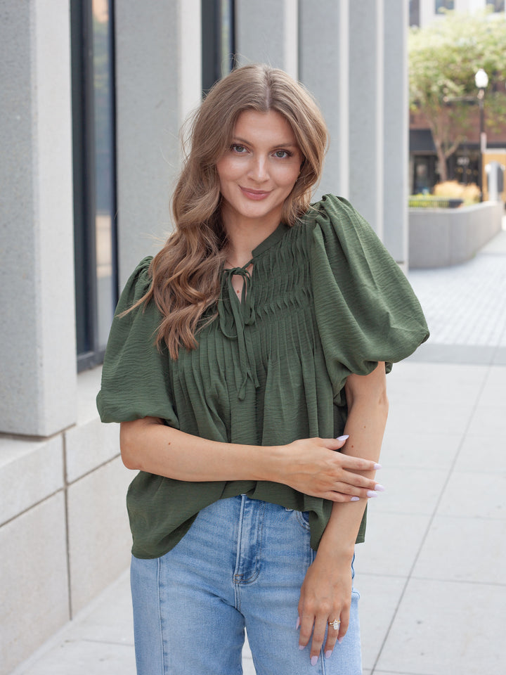 A woman wearing an olive green puff-sleeve blouse with a smocked neckline and tie-front keyhole detail, paired with light-wash blue jeans, standing outdoors on a paved walkway.