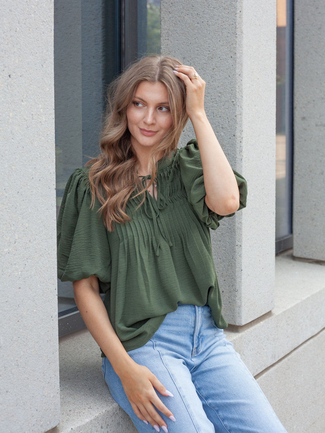 A woman wearing an olive green short-sleeve blouse with puff sleeves and a smocked bodice, paired with light blue jeans, sitting on concrete steps.