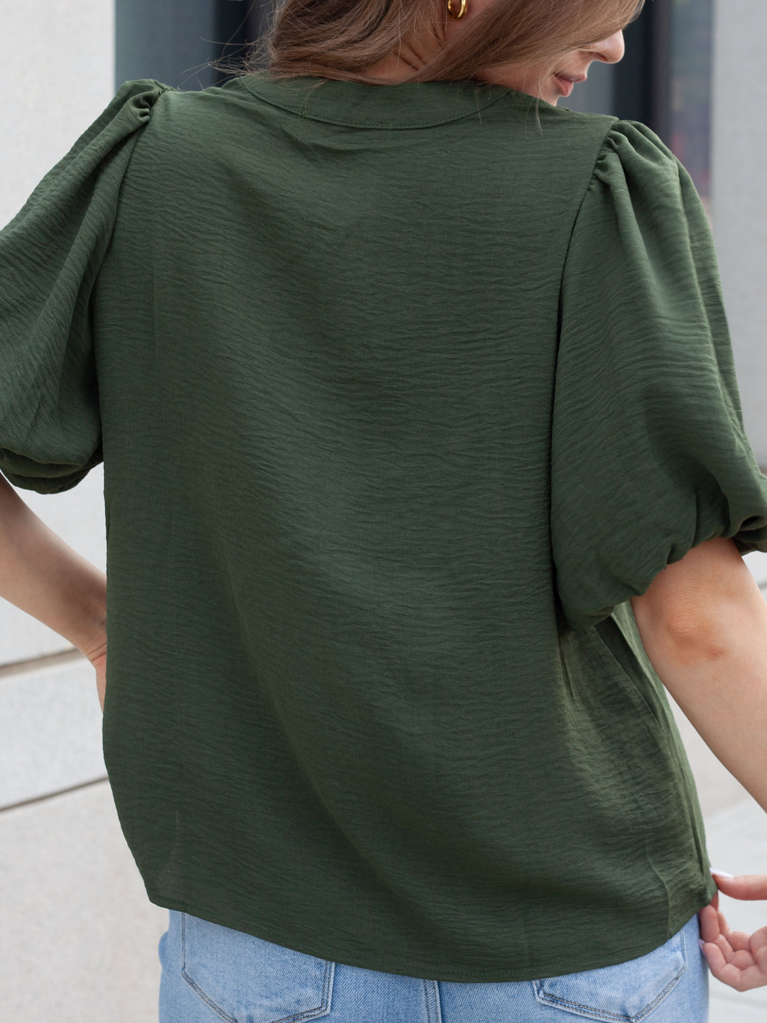 A woman wearing an olive green short-sleeve blouse with voluminous puff sleeves, shown from the back. The blouse has a textured crinkle fabric and is paired with light blue jeans.