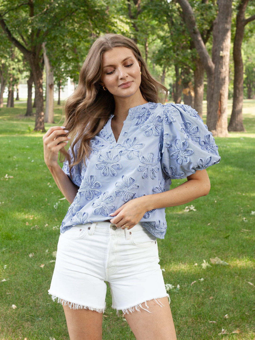 A woman standing outdoors in a park wearing a light blue floral puff-sleeve blouse and white denim shorts.