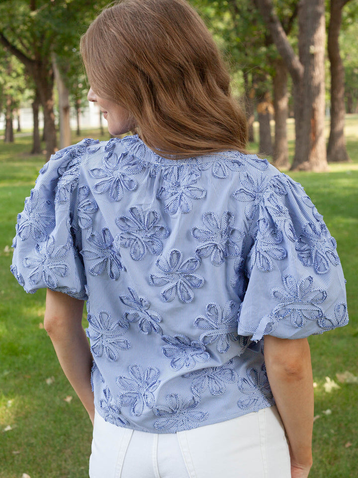 A woman viewed from behind wearing a light blue puff-sleeve blouse with raised floral embroidery, paired with white pants, standing outdoors in a park with trees and grass in the background.