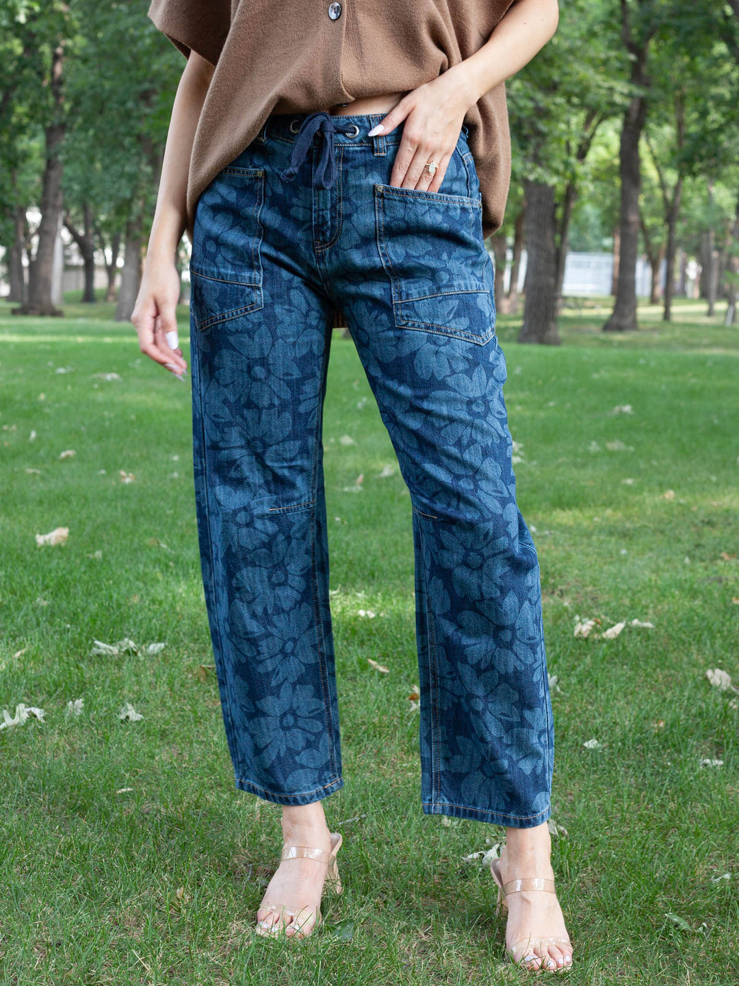 A person standing on a grassy park lawn wearing blue patterned wide-leg jeans, a brown short-sleeve top, and clear high-heeled sandals.