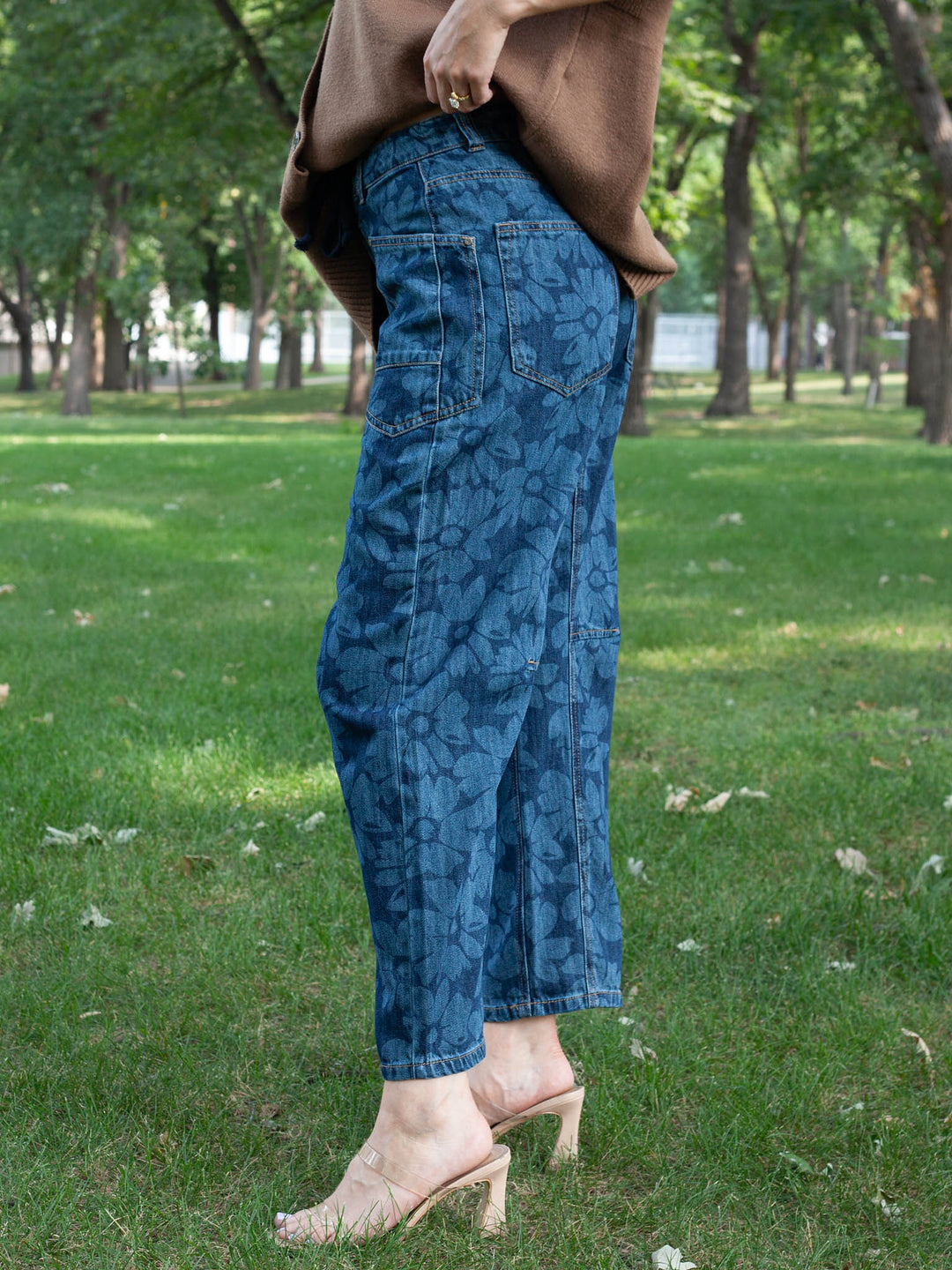 A person standing outdoors on grass wearing blue patterned cropped jeans, a brown top, and beige heeled sandals.
