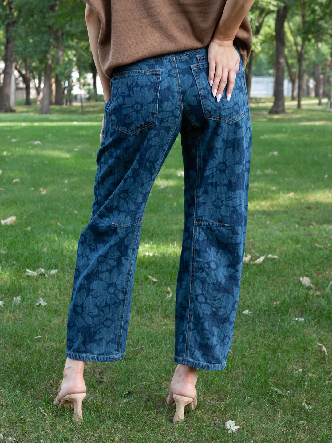A person standing outdoors on grass wearing blue floral-print jeans, a brown sweater, and beige high heels, shown from the back.