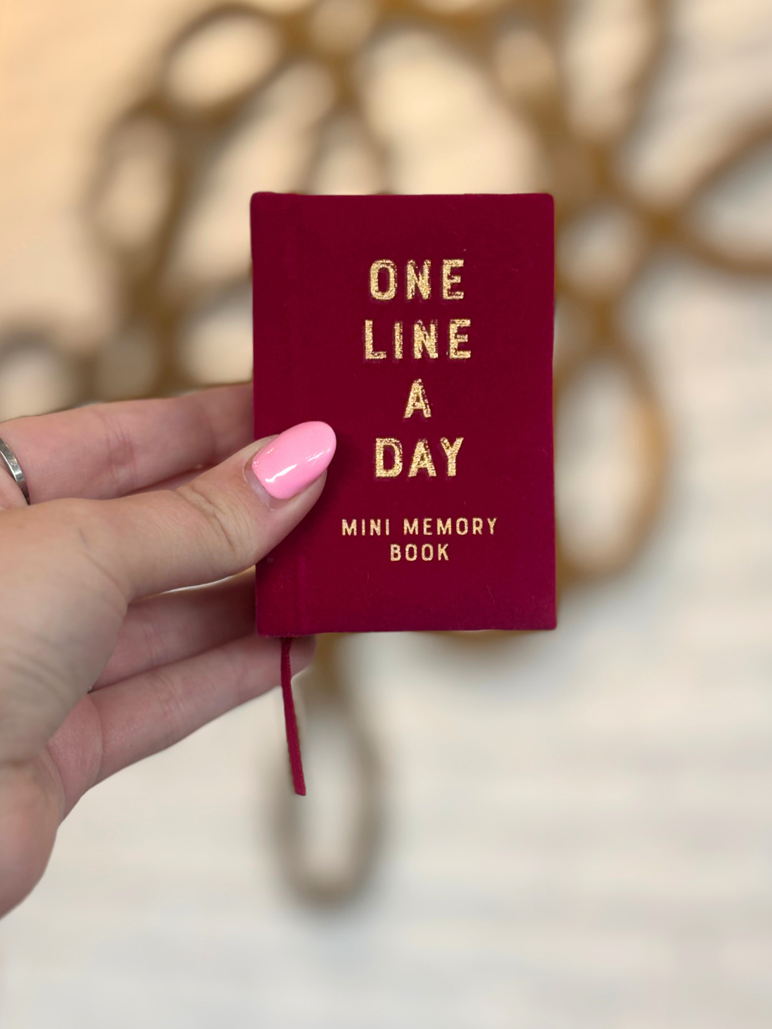 A hand with pink nail polish holds a small burgundy mini memory book with gold foil text that reads &