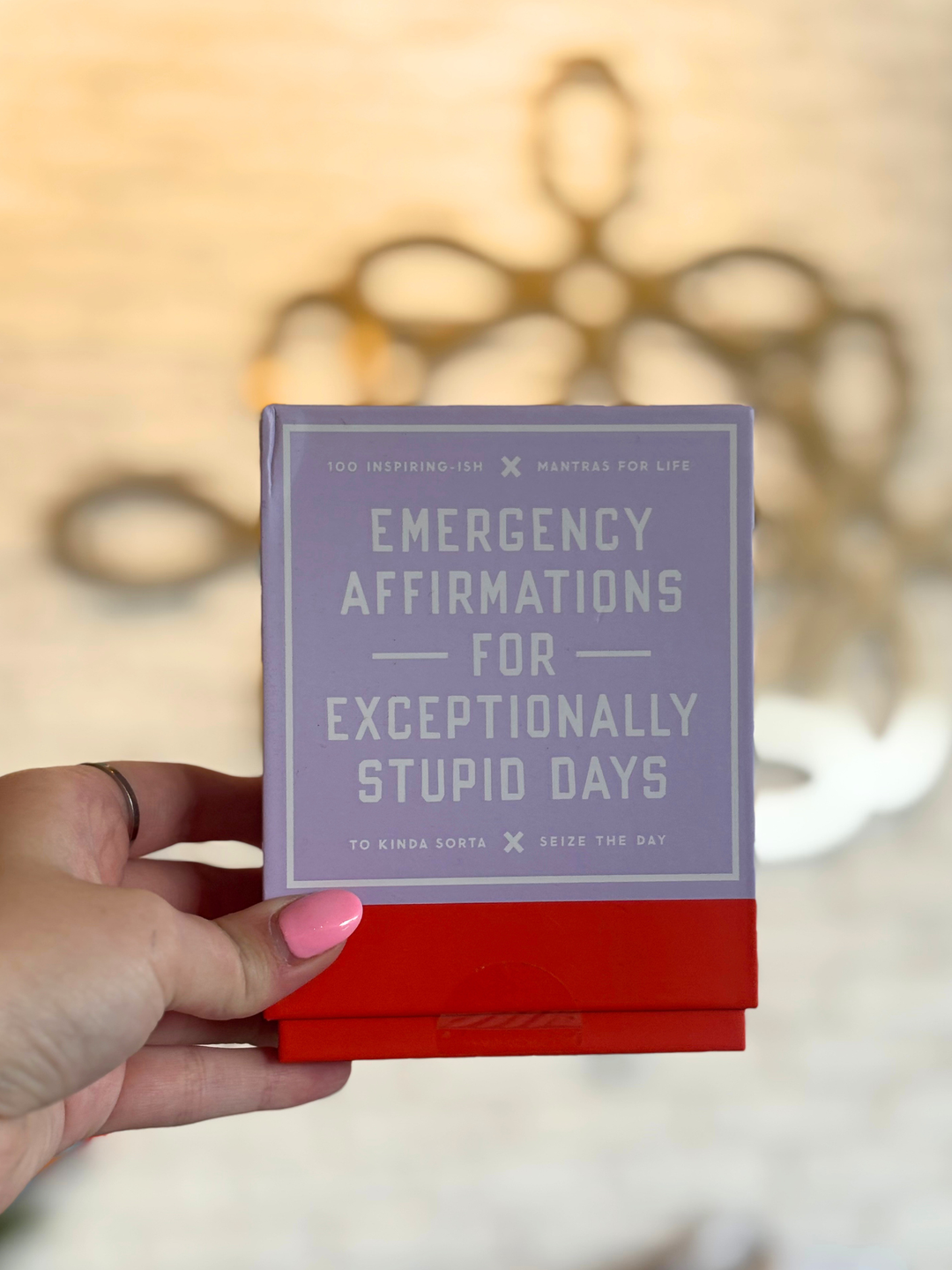 A hand with pink nail polish holds a small square card or box featuring the text &