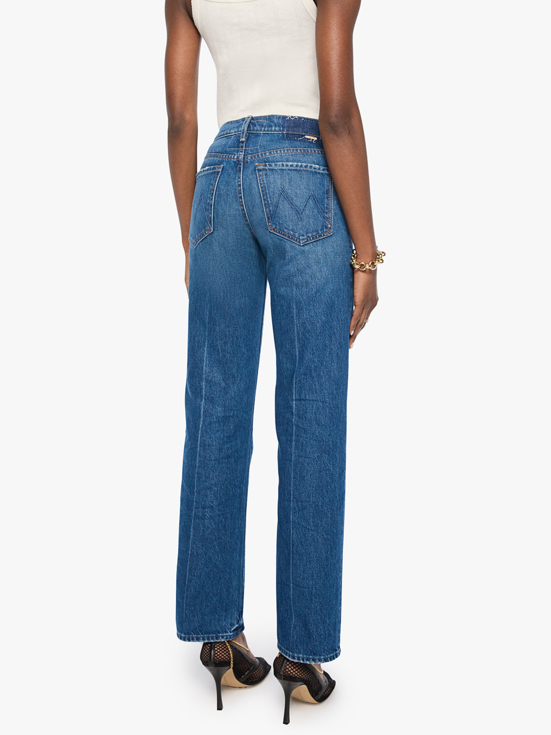 A model shown from the back wearing high-waisted, wide-leg blue denim jeans, a white ribbed tank top, and black high-heeled sandals against a plain white background.
