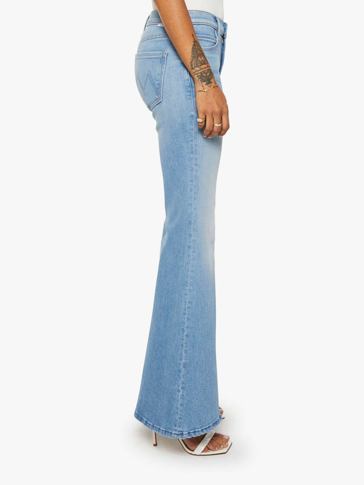 A side profile view of a model wearing light-wash blue flare jeans and white heeled sandals against a plain white background.