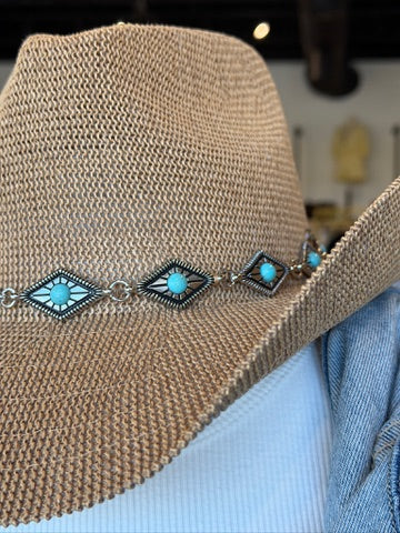 A close-up of a woven straw cowboy hat with a silver-toned band featuring turquoise and geometric inlay details, displayed on a mannequin head with a light blue shirt underneath.