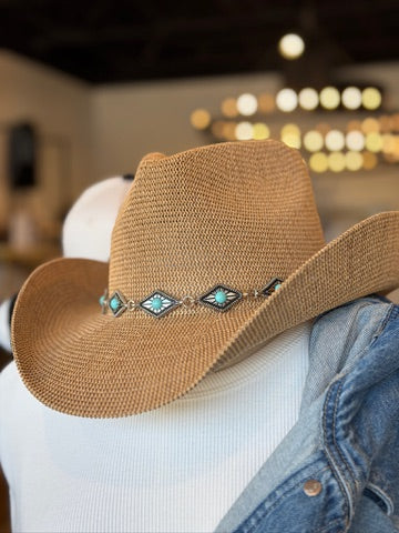 A tan woven cowboy hat with turquoise and silver geometric band embellishments, displayed on a white mannequin torso with a blue denim shirt underneath in a softly blurred indoor setting.
