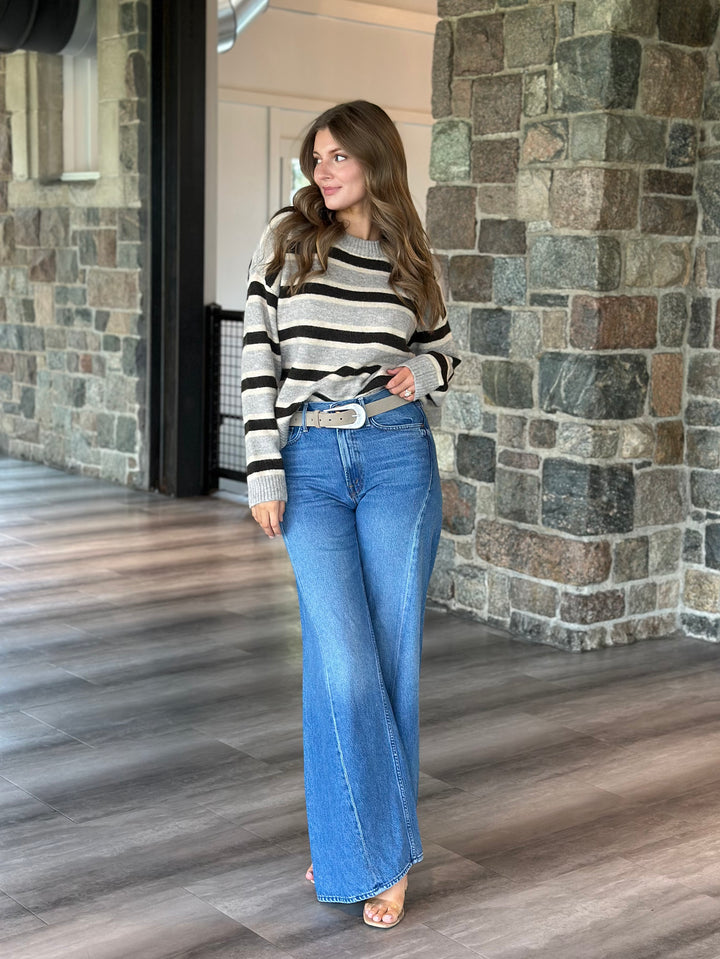 A woman modeling a black and white striped long-sleeve knit top with wide-leg blue jeans and a silver belt, standing in a stone-walled indoor hallway.