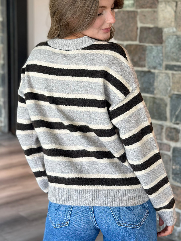 A woman wearing a gray knit sweater with horizontal black and cream stripes, paired with blue jeans, shown from a side-back angle against a stone wall background.