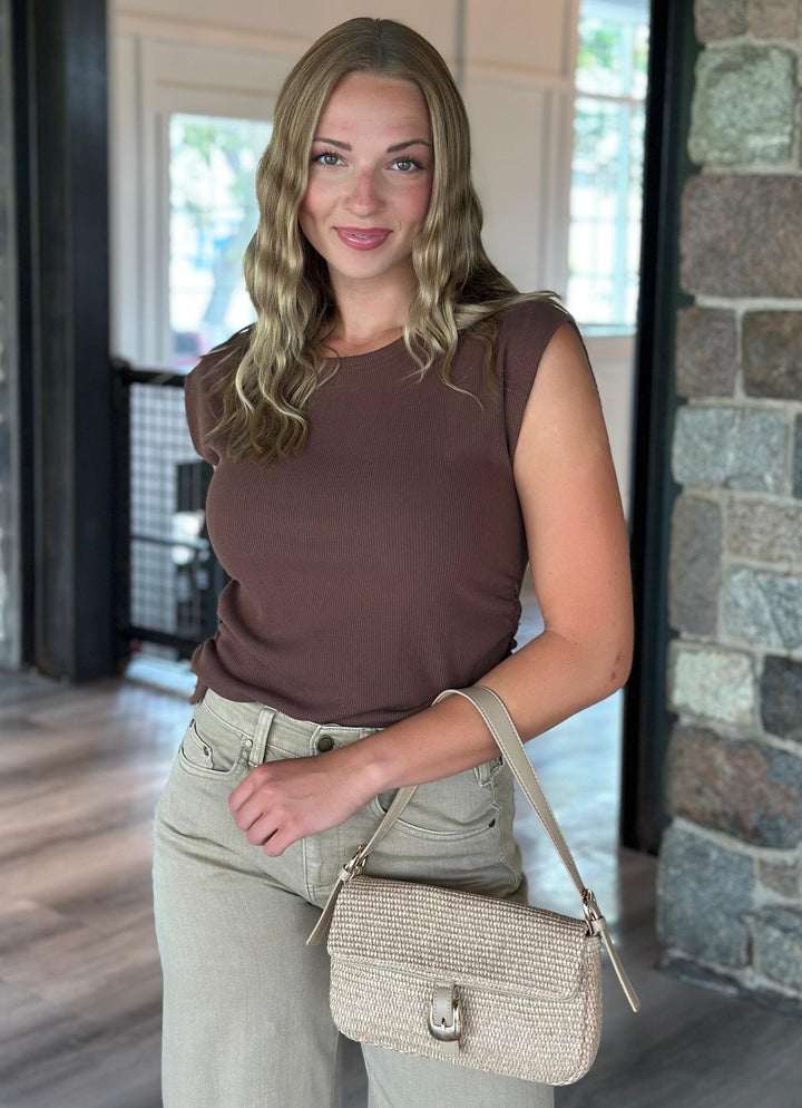 A woman wearing a brown sleeveless top and beige pants, holding a beige woven shoulder bag in a modern indoor setting with a stone pillar.