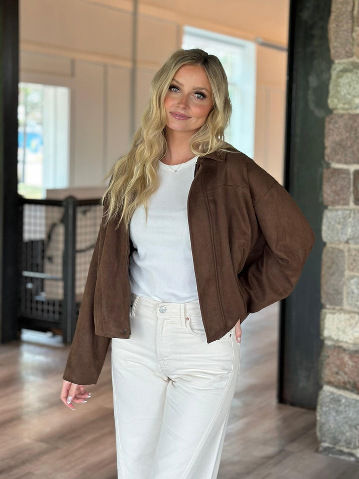 A woman modeling a brown suede-like cropped jacket over a white top and white pants, standing in an indoor setting with a stone column and wood floor.