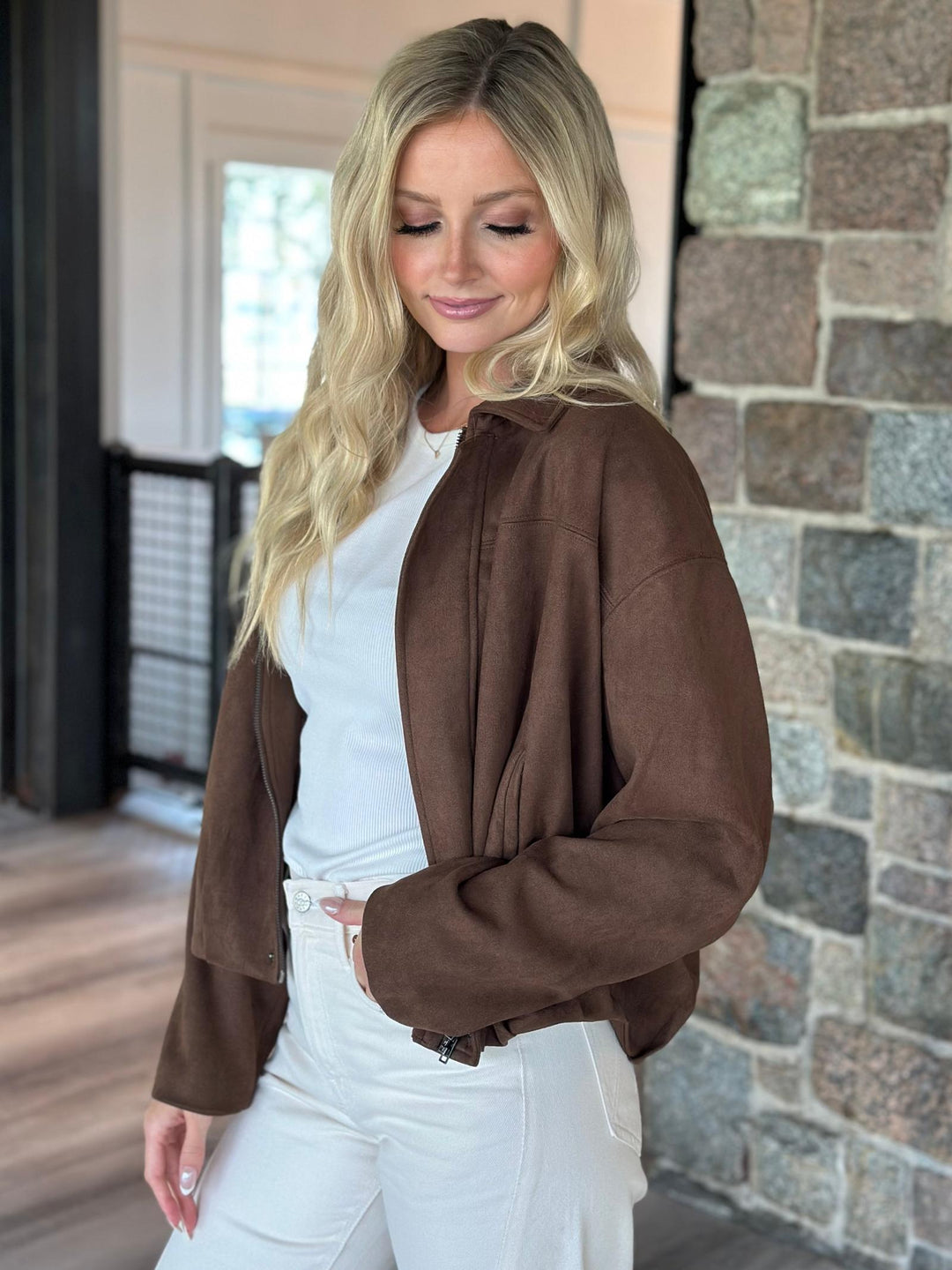 A woman wearing a brown suede-like cropped jacket over a white top and white pants, standing indoors next to a stone wall.